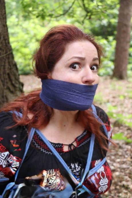 Pale redhead is silenced with a gag while tied up in her clothes under a tree 35774830